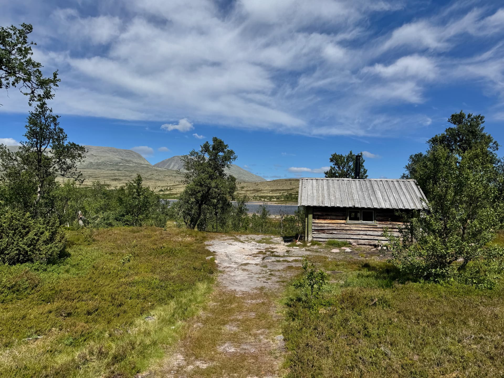 Weathered log rescue hut with a metal chimney beside the trail near Granåsen, mountains and a lake in the background