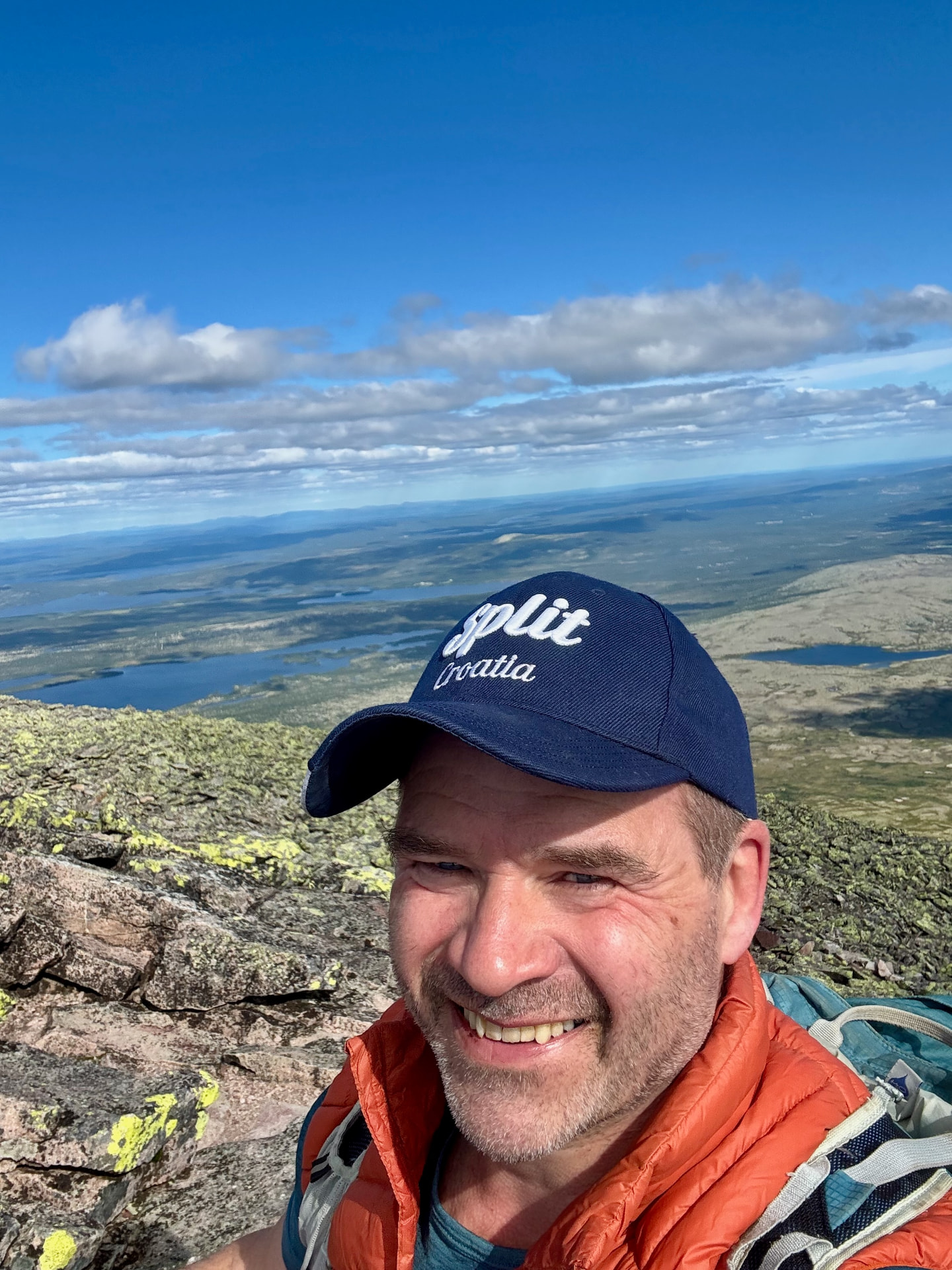 Smiling hiker in an orange down jacket and dark cap at the summit of Sølen, lakes and forest far below